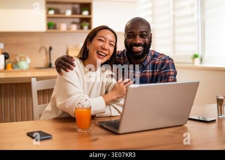 Fröhliches erwachsenes Paar sitzt zusammen an einem Holztisch und benutzt Laptop für Videokommunikation. Sie lächeln und schauen in die Kamera. Stockfoto