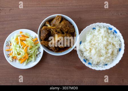 Authentische südasiatische Mahlzeit mit gedämpftem weißem Reis, scharfem Entencurry und frischem Gemüsesalat, serviert in weißen Schüsseln auf einem Holztisch. Draufsicht eines Stockfoto