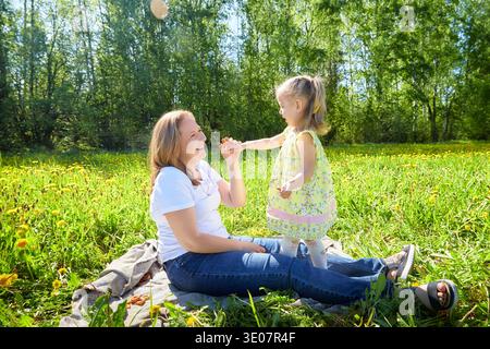 Mutter und Tochter genießen einen verspielten Moment in einem üppig grünen Feld mit gelben Blumen unter hellem Sonnenlicht. Stockfoto