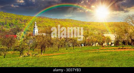 Sonnenuntergang über bergiger Landschaft im Frühling. Wunderbares Wetter mit bewölktem Himmel und Regenbogen. Ländlicher Obstgarten am Rande des Dorfes. Stockfoto