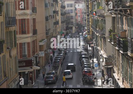 Steile Straße in Nizza, Frankreich, mit mehrstöckigen Gebäuden mit kunstvollen Balkonen und bunten Rollläden, voll mit geparkten Fahrzeugen. Stockfoto