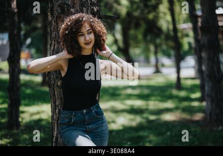 Junge Frau in schwarzem ärmellosem Oberteil lehnt sich im Park an einen Baum und genießt einen sonnigen Tag im Freien Stockfoto