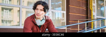 Ein junger Mann mit lockigen Haaren entspannt sich draußen an einem Frühlingstag, gekleidet in einer Sportjacke, einem Banner Stockfoto