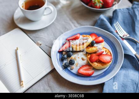 Blick von oben auf hausgemachte Keto Cottage Käse Pfannkuchen mit frischen Erdbeeren und Heidelbeeren, eine Schüssel Erdbeeren und eine Tasse Tee auf einem Tisch mit offenem Notizbuch und Stift Stockfoto