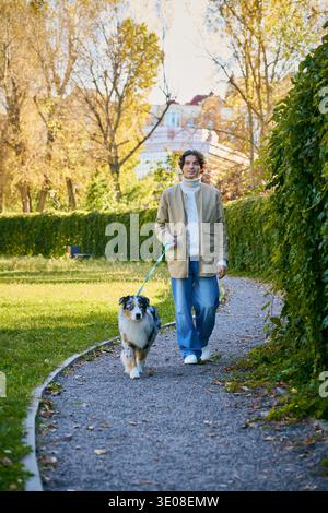 Ein junger Mann mit lockigen Haaren läuft zufrieden neben seinem Hund des Australian Shepherd in einem lebhaften Park. Stockfoto
