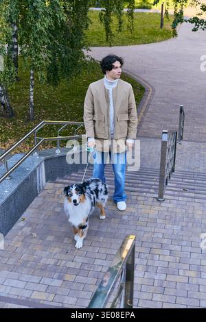Ein junger Mann schlendert mit seinem verspielten australischen Schäferhund in Frühlingssonne die Treppe hinunter. Stockfoto