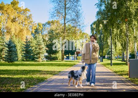 Ein junger Mann in einer Frühlingsjacke schlendert glücklich mit seinem australischen Schäferhund in einem lebhaften Park. Stockfoto