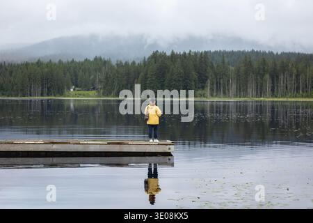 Weibliche Touristen, die morgens auf einem hölzernen Pier in einer natürlichen Parklandschaft stehen. Rückansicht einer jungen Frau in gelber Jacke, die auf einem Steg im Vorsprung steht Stockfoto