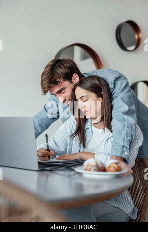 Ein junges Paar genießt einen Morgen zu Hause und lächelt, während es zusammen an einem Laptop arbeitet. Das warme Licht und die Teamarbeit reflektieren Liebe, Produktivität und eine modische Note Stockfoto