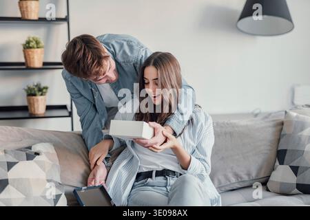 Aufgeregter junger kaukasischer Mann, der aufgeregten Freundin ein Überraschungsgeschenk gibt, zurücksteht, Hand auf Augen legt, lacht. Fröhliches Millennial-Paar lacht Stockfoto