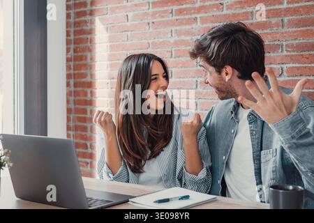 Überglücktes Millennial-Paar sitzen am Schreibtisch, schauen auf den Laptop-Bildschirm und fühlen sich euphorisch mit Online-Gewinn, aufgeregt, glücklich, junger Mann und Frau, lesen gute Nachrichten, hören Sie Stockfoto