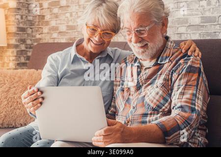 Süße alte Leute, die auf dem Sofa sitzen und ihren Laptop zusammen zum Einkaufen und Surfen im Internet benutzen. Zwei Erwachsene, die eine Brille tragen Stockfoto