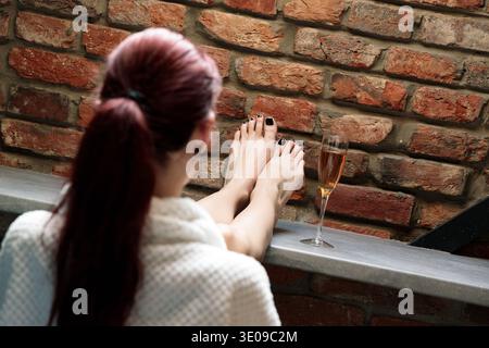 Eine Frau entspannt sich mit den Füßen auf einer Theke, ein Glas Champagner neben ihr, vor einem rustikalen Backsteinmauerhintergrund Stockfoto