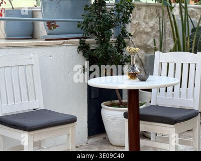 Sitzbereich im Café im Freien mit zwei weißen Stühlen und einem Tisch mit Pflanzen im Hintergrund. Mobiles Foto Stockfoto