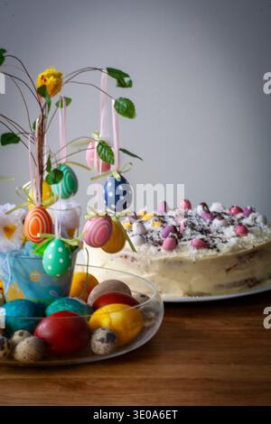 Festlicher Osterkuchen mit weißer Zuckerguss, dekoriert mit Mini-Schokoladeneiern und Schokoladenspänen, auf einem Tisch mit bunten hängenden Eiern Stockfoto