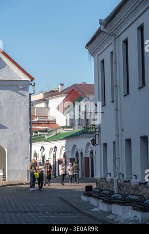 Minsk, Weißrussland - 8. März 2026: Fußgänger, die entlang der historischen Straße mit weißen Gebäuden und der belarussischen Flagge in der Altstadt von Minsk spazieren. Städtisch Stockfoto