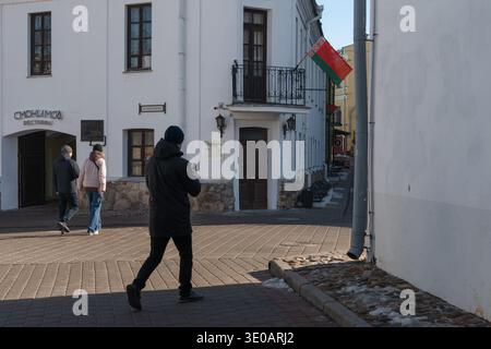 Minsk, Weißrussland - 8. März 2026: Fußgänger entlang der historischen Straße mit weißen Gebäuden, weißrussische Flagge an der Fassade im Zentrum von Minsk. Stadtstraße Stockfoto