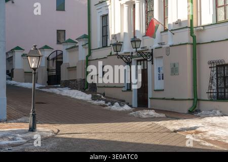 Minsk, Weißrussland - 8. März 2026: Historische Gebäudefassade mit belarussischer Flagge, Eingangstor, Straßenlaternen und Kopfsteinpflasterstraße Stockfoto