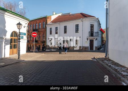 Minsk, Weißrussland - 8. März 2026: Historische Straße mit weißem Eckgebäude, weißrussischer Flagge, Fußgängern und Kopfsteinpflaster im alten Stadtteil von Stockfoto