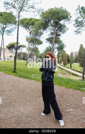 Junge rothaarige Frau mit Sonnenbrille im grünen Stadtpark mit Kiefern. Reise-Lifestyle und urbane Naturszene. Stockfoto