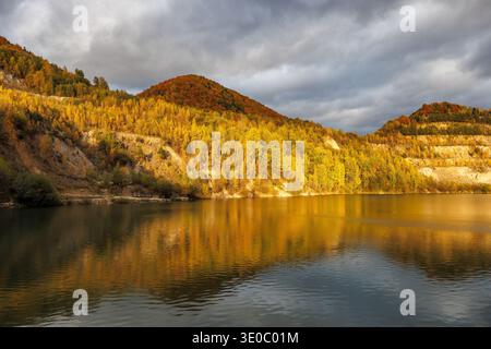 Ein ruhiger See reflektiert die goldenen Farben des Herbstblatts auf den umliegenden Hügeln und strahlt ein warmes Leuchten unter einem bewölkten Himmel aus. Überfluteter Steinbruch in der Nähe des Stockfoto