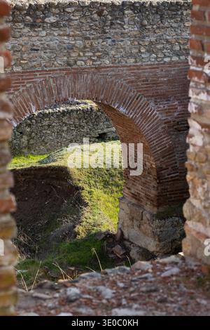Blick auf den alten römischen Backsteinbogen durch Steinruinen mit sonnendurchflutetem grünem Gras und bröckelnden Mauern aus Mauerwerk, historische architektonische Texturen in natürlicher Natur Stockfoto