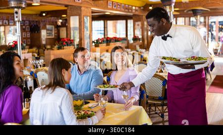 Ein afroamerikanischer Kellner serviert Speisen für eine fröhliche Gruppe von Erwachsenen im Restaurant Stockfoto