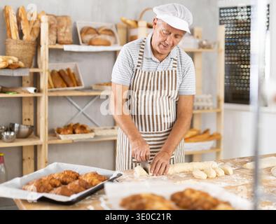 Senior man arbeitet in der Bäckerei als Bäcker, schneidet Teig in Portionen, formt Teigstücke, um Brötchen zu kreieren. Stockfoto