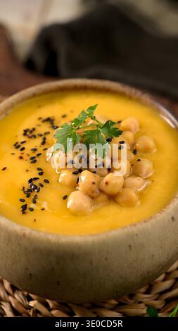 Eine lebendige Schüssel mit samtiger Kürbis- oder Kürbissuppe, serviert in einer rustikalen Keramikschale. Garniert mit proteinreichen Kichererbsen und frischer Petersilie unter warmen, Stockfoto