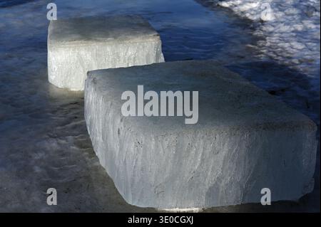 Zwei große rechteckige Blöcke aus klarem Eis auf einer gefrorenen Küste, beleuchtet von kalter Wintersonne mit umliegendem Schnee und dunkelblauem Eis c Stockfoto