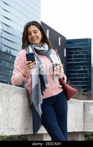 Frau mittleren Alters, die sich auf das Geländer auf dem büroplatz stützt, Telefon benutzt, Kaffee hält, rote Tasche trägt Stockfoto