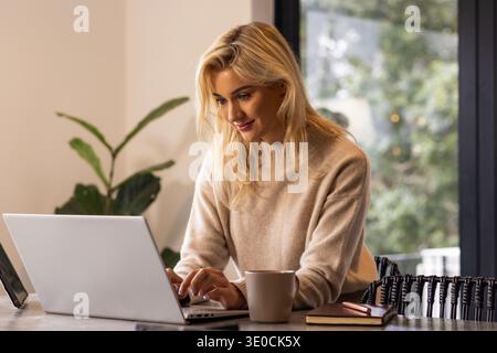 Frau in 20ern, die einen beigefarbenen Pullover trägt, arbeitet an silbernem Laptop am Tisch mit Kaffeetasse, Pflanze Stockfoto