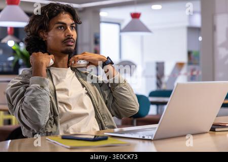 Indischer Mann, der am Coworking-Tisch sitzt und weiße Kopfhörer mit offenem Laptop in khakifarbener Jacke anpasst Stockfoto