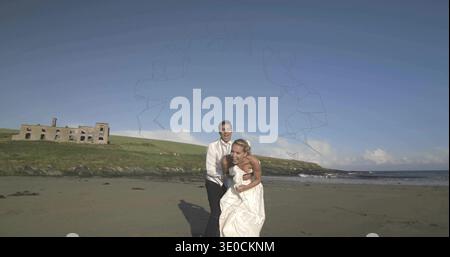 Spazieren Sie Braut und Bräutigam, umarmen Sie sich und bewegen Sie sich vorwärts am Sandstrand, zeigen Sie Hochzeitskleid und Ruine Stockfoto