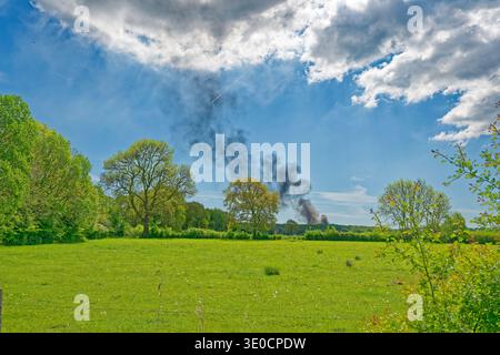 Ein üppiges grünes Feld mit verstreuten Bäumen und schwarzem Rauch, der in der Ferne unter einem teilweise bewölkten blauen Himmel an einem hellen Tag aufsteigt. Stockfoto