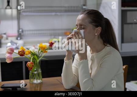 Seoul, Südkorea - 03.12.2026: Junge Frau, die ein starkes Niesen erlebt, während sie ein Tuch zu Hause in der Nähe der Frühlingsblumen hält Stockfoto