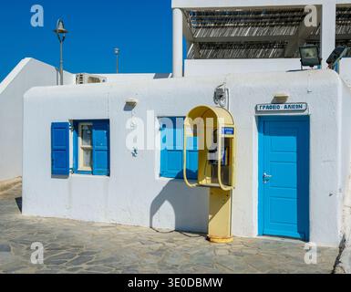 01.09.2025, Griechenland, Mykonos, Mykonos - alte gelbe Telefonzelle vor einem weißen Haus mit blauen Fensterläden in den alten Straßen der Stadt Mykonos. Stockfoto