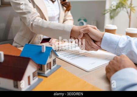 Ein Immobilienmakler und ein Kunde schütteln sich die Hände, nachdem sie einen Kaufvertrag unterzeichnet haben. Der Schreibtisch zeigt Modellhäuser, einen Vertrag und Kaffeetassen, Stockfoto