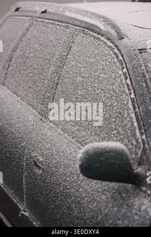 Das Auto ist an einem kalten Wintermorgen mit Frost und leichtem Schnee bedeckt. Gefrorene Fenster und Seitenspiegel veranschaulichen raue Witterungsbedingungen, Wintertranspo Stockfoto