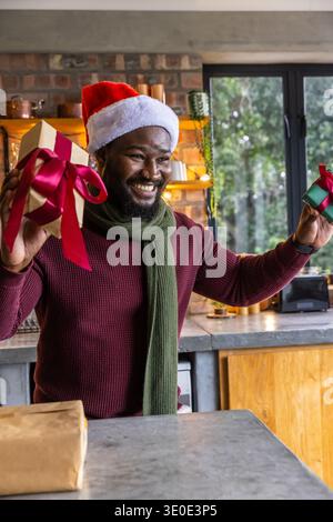 Afroamerikaner mit Weihnachtsmütze und grünem Schal, der zwei Geschenke in der Küche hält Stockfoto