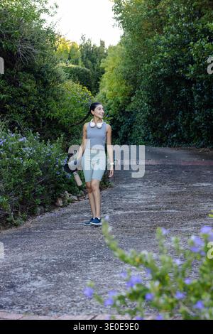 Asiatische Frau, die auf einem gepflasterten Parkweg in Sportbekleidung läuft, mit gerollter Yogamatte und Wasserflasche. Outdoor, Fitness, Trail, Grün, ruhig, athleisure, Stockfoto