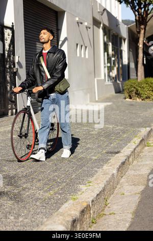 Mann in den Zwanzigern, der Rennrad mit roter Felge auf dem Bürgersteig neben der Ladenfront läuft und eine Oliventasche hält. Fahrrad, Radfahrer, Urban, Stadtlandschaft, Crossbody, lässig, Su Stockfoto