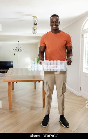 Mittelalter afroamerikanischer Mann, der zu Hause im Essbereich steht und eine GLASKISTE mit Flaschen hält. Recycling, nachhaltig, modern, hell, interio Stockfoto