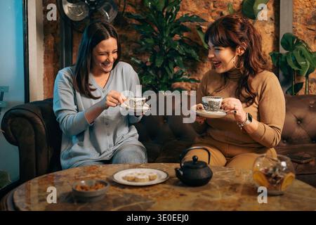Zwei fröhliche junge Freundinnen sitzen auf einem Ledersofa in einem Café, lachen und genießen Tee mit Snacks auf einem Marmortisch, die au repräsentieren Stockfoto