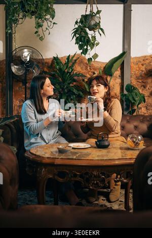 Zwei junge Freundinnen sitzen auf einem braunen Ledersofa in einem gemütlichen urbanen Café, trinken Tee und unterhalten sich, während sie von grünen Pflanzen umgeben und rustikal sind Stockfoto