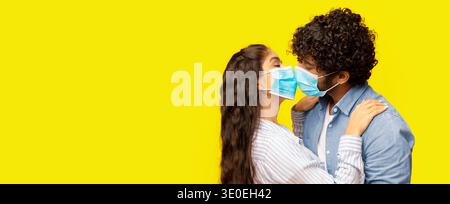 Ein Paar teilt einen Kuss, während es Masken vor einem hellgelben Hintergrund trägt Stockfoto