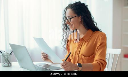 Frau arbeitet an Dokumenten, während sie im hellen Raum mit Laptop und Stift am Schreibtisch sitzt Stockfoto