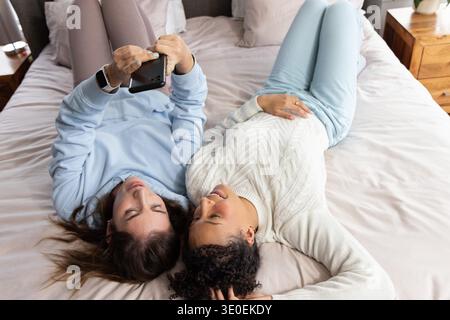 Verschiedene Freundinnen liegen nebeneinander auf dem Bett, halten das Telefon oben, zeigen Smartwatch und Klingeln. Gerät, Damen, Freizeit, gemütlich, Loungewear, Schlafzimmer, neutral || Model freigegeben Stockfoto