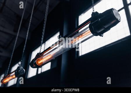 Reihe von industriellen Glühlampen. Eine Niedrigwinkelaufnahme von zwei Hängeleuchten aus röhrenförmigem Filament im Vintage-Stil, die in einer Reihe in einem dunklen Lager hängen. Stockfoto