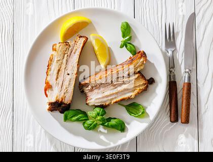 Gebratene Schweinebauchrippchen auf weißem Teller, serviert mit frischen Zitronenscheiben, Basilikumblättern und rustikalem Besteck auf weißem Holztisch, horizontale Sicht, flache Lage, Stockfoto